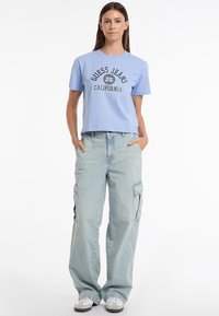 Šviesiai mėlyna medvilninė marškinė su "Guess Jeans California" grafika, derinama su šviesiai mėlynomis kargo kelnėmis, turinčiomis šonines kišenes ir laisvu apipavidalinimu.