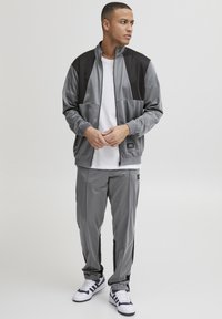 Grå idrettsjakke med svarte detaljer; zip-up jakke og bukser med elastisk midje; glatt, lett stoff; kombinert med hvite sneakers.