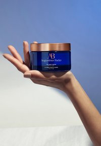Blå glasbeholder med Augustinus Bader body cream og et bronze låg. Etiketten har gyldne bogstaver. Holdes i en hånd mod en lys baggrund.