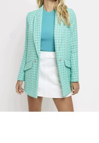 Blazer a scacchi turchesi con revers, tasche frontali e dettagli decorativi. Indossato sopra un top a coste turchese e una gonna bianca.