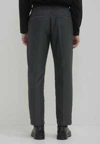 Pantalons tissés gris avec poches latérales et arrière. Taille élastique, coupe fuselée et chevilles ourlées. Portés avec des chaussures noires.