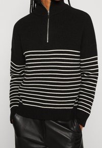 Schwarzer gestrickter Pullover mit hohem Kragen und horizontalen weißen Streifen. Verfügt über einen Reißverschluss vorne sowie eine gerippte Struktur am Saum und an den Bündchen.