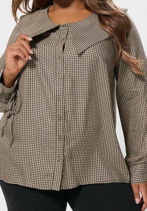 Femme portant une chemise à carreaux marron et noire à boutons avec un large col, tenant une partie du col avec la main gauche.
