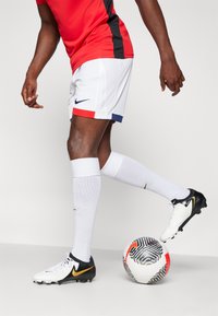 Rode en zwarte Nike jersey in combinatie met witte shorts met een marineblauw accent. Speler draagt witte sokken en zwart/witte voetbalschoenen met een gouden swoosh.