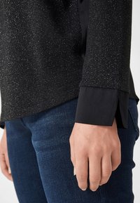 Schwarzes Shirt mit glitzernder Textur am oberen Teil und einfarbigen schwarzen Ärmeln. Der Bund hat eine weit geschnittene Öffnung. Getragen mit dunkelblauen Jeans.