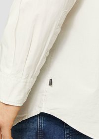 Chemise à boutons créme légère avec un tissu texturé, un poignet replié et une petite étiquette de marque cousue sur le côté ; portée sur un jean en denim bleu.