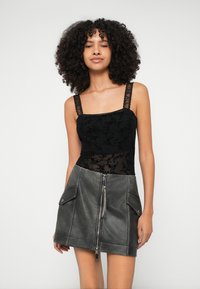 Mulher com cabelo encaracolado vestindo um top sem mangas de renda preta com flores e uma saia de couro cinza escuro com zíper frontal e bolsos com aba.