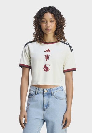 Ung kvinde med krøllet hår iført en hvid kort sportstrøje med røde og sorte logoer, kombineret med lyseblå højttaljede jeans.