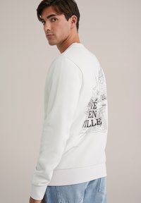 White sweatshirt featuring a graphic design on the back, depicting a cityscape in black outlines with text "EN VILLE" prominently displayed.