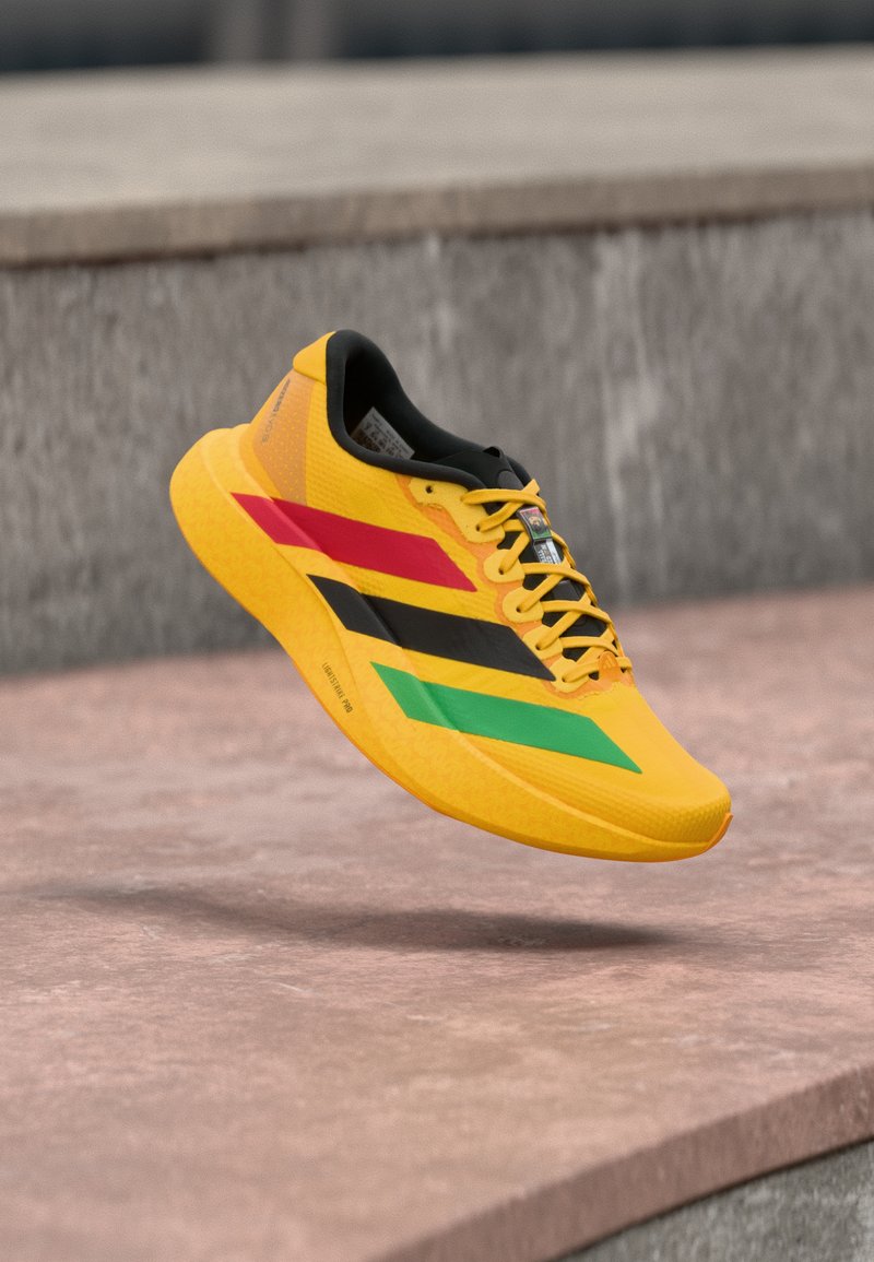 Chaussure de course jaune avec des rayures rouges, noires et vertes, doublure intérieure noire et lacets jaunes, flottant au-dessus d'une surface en pierre en plein air.