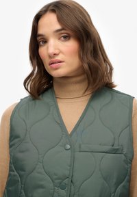 Zielona kamizelka pikowana z dekoltem w kształcie litery V, zatrzaskami i kieszenią; noszona na beżowym golfie, prezentująca gładkie faktury i kontrasty.
