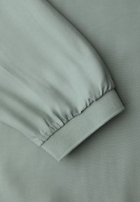 Tissu vert clair à texture lisse, doté d'un poignet de manche froncé et d'un bord plat au bas, complétant le design global.