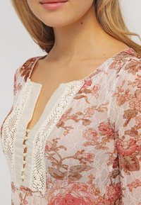 Top in pizzo floreale con scollatura testurizzata color crema e dettagli con bottoni, motivi floreali rosa morbido e marrone, e un design delicato.