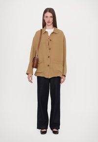 Brown oversized button-up jacket with two front pockets, worn over a white shirt and dark wide-leg jeans; brown shoulder bag.