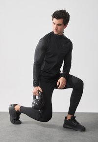 Conjunto atlético negro que incluye una parte superior ajustada de manga larga y leggings. De rodillas sosteniendo una pesa rusa negra de 10 kg.