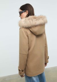 Donna che indossa un cappotto di lana beige con cappuccio bordato di pelliccia, occhiali da sole tartaruga e jeans azzurri chiari, di spalle su uno sfondo neutro.