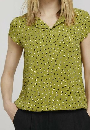 Femme portant un chemisier vert olive à manches courtes avec un petit motif abstrait noir et blanc et un col, associé à un pantalon noir.