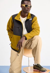 Giacca in pile e nylon di colore nero e senape, con tasche zippate, camicia beige, pantaloni khaki e sneaker olive con accenti rosa.