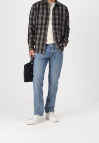 Homme portant une chemise à carreaux noir et blanc, un t-shirt blanc, un jean bleu, des baskets blanches, tenant un petit sac noir d'une main.