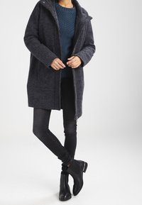 Abrigo de lana gris oscuro con cierre de cremallera, dos bolsillos frontales y un alto cuello, combinado con un suéter azul texturizado y jeans ajustados negros.