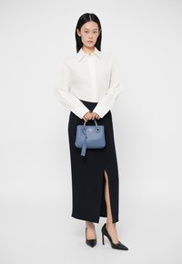 Λευκό πουκάμισο με κουμπιά, μαύρη μακριά φούστα με σκίσιμο, μαύρα ψηλά τακούνια, κρατώντας μια μικρή μπλε δερμάτινη τσάντα με ετικέτα. Καθαρός, μοντέρνος σχεδιασμός.