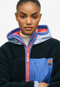 Jonge vrouw met donker haar, gekleed in een zwart en blauwe fleecjas met een blauw en koraalkleurige capuchon, dichtgeritst over een beige coltrui.