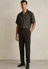 Camisa de cuadros de manga corta en tonos oscuros con un cuello relajado, combinada con pantalones negros a medida y zapatos de cordones negros.
