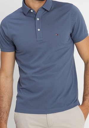 Mann trägt ein eng anliegendes blaues Polohemd mit kurzen Ärmeln, Knöpfen und kleinem rechteckigem Logo auf der Brust, kombiniert mit hellbeigen Hosen.