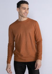 Maglione lavorato a maglia color ruggine con colletto rotondo, polsini a coste e righe texturate sul tessuto. Indossato con jeans scuri.