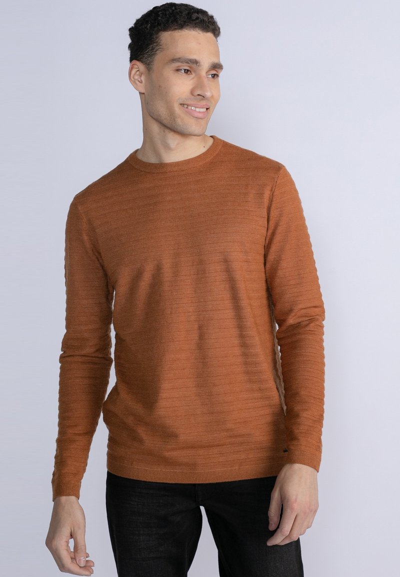 Maglione lavorato a maglia color ruggine con colletto rotondo, polsini a coste e righe texturate sul tessuto. Indossato con jeans scuri.