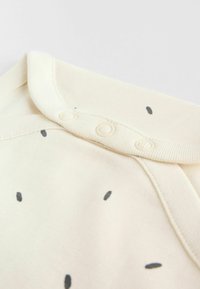 Vauvan vaalea kerrospuku, jossa on ribbareunus ja neljän napin kiinnitys, ja kankaassa on hienovaraisia mustia pilkkuja.