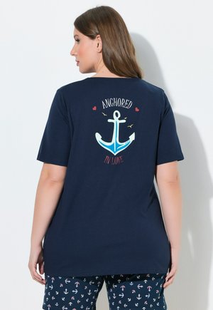 Frau mit langen braunen Haaren, die ein dunkelblaues T-Shirt mit Anker-Motiv und dem Text "Verankert in Liebe" trägt, kombiniert mit Shorts mit Anker-Muster.
