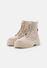 Bottines en daim beige avec une semelle plateforme épaisse, fermeture à lacets à l'avant et détail zippé. Finition texturée et surpiqûres apparentes.