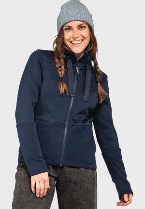 Marineblaue Fleecejacke mit Reißverschluss, strukturiertem Oberteil, einfarbigem unterem Abschnitt und verstellbarem Kapuzenbereich. Kombiniert mit dunklen Hosen und einer gestrickten Mütze.
