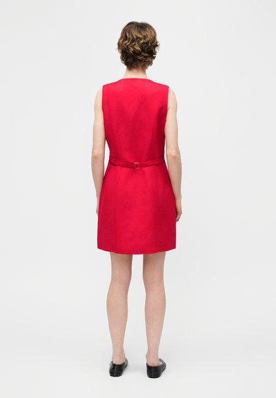 Άτομο με κοντά καστανά μαλλιά, ντυμένο με αμάνικο κόκκινο φόρεμα με ζώνη, στέκεται πλάτη σε απλό ανοιχτό φόντο.