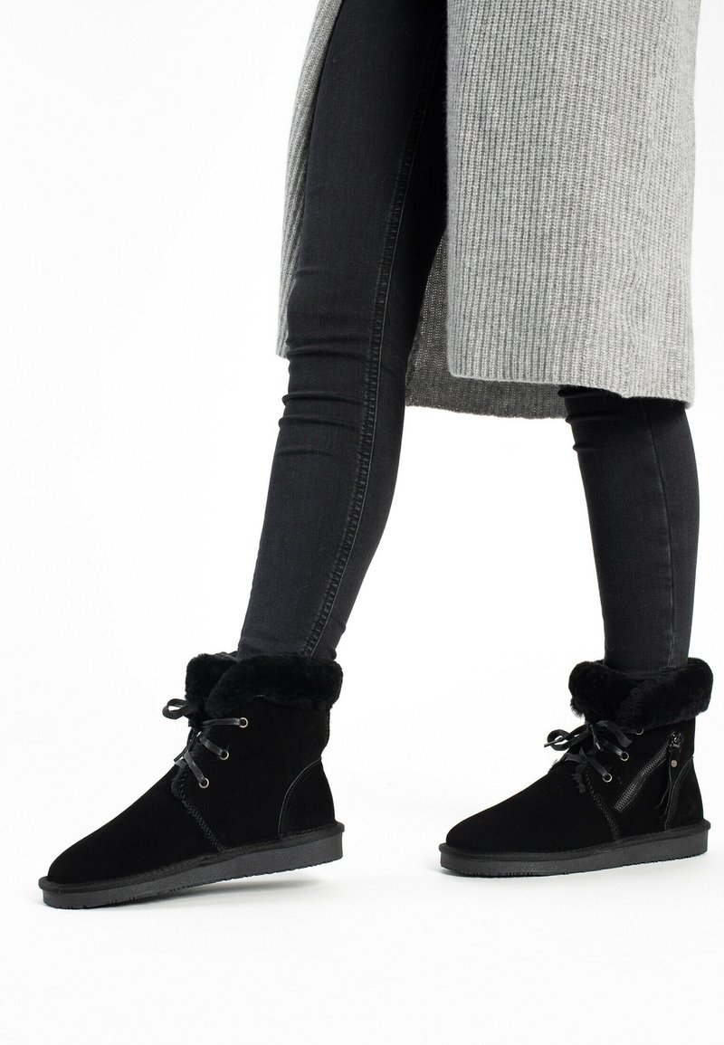 Bottes montantes en cuir noir doublées de fourrure, avec lacets et fermeture éclair sur le côté, portées par-dessus un jean skinny noir et un manteau long gris en maille côtelée.
