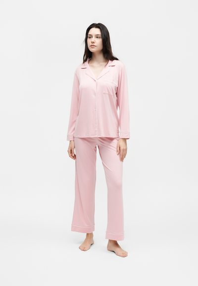 Γυναίκα στέκεται ξυπόλυτη φορώντας φωτεινό ροζ πυτζάμα με κουμπιά και παντελόνια με φαρδιά μπατζάκια σε λευκό φόντο.