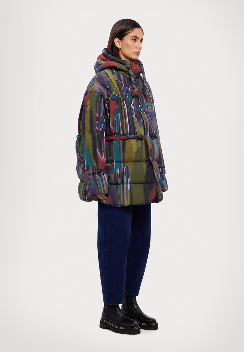 Veste en duvet avec un design abstrait multicolore, dotée d'une capuche, de poches avant et d'une coupe ample. Associée à un jean bleu foncé et des bottes noires épaisses.