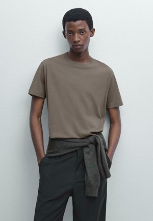 Braunes Rundhals-T-Shirt aus weichem Stoff, kombiniert mit dunklen Hosen. Ein grauer Pullover ist um die Taille gebunden. Einfacher, lässiger Look.