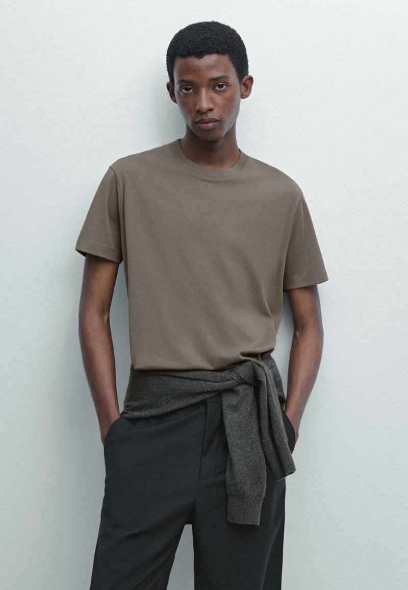 Hnědé tričko s kulatým výstřihem vyrobené z měkké tkaniny, v kombinaci s tmavými kalhotami. Šedý svetr je uvázaný kolem pasu. Jednoduchý, ležérní vzhled.