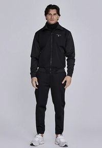 Čierna atletická tepláková súprava z hladkej tkaniny, ktorá obsahuje bundu na zips a zúžené nohavice. Zlaté logo na bunde.