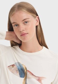White long-sleeve t-shirt featuring a graphic of a fish with blue and blonde accents on one side. Soft fabric with a round neckline.