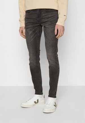 Mand iført slim-fit falmede sorte jeans, hvide sneakers med grønne logoer og en beige sweatshirt med et lille flaglogo på ærmet.