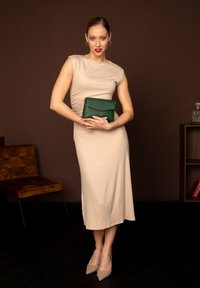 Béžové přiléhavé šaty s krátkými rukávy, zelená texturovaná kabelka a nahé špičaté podpatky. Hnědý pozadí a dřevěná židle v scéně.