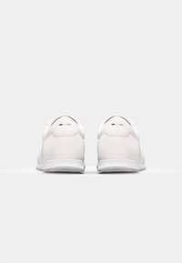 Chaussures de sport blanches avec tige en cuir lisse, col rembourré, semelle texturée, et accents minimalistes noirs, blancs et bruns au talon.