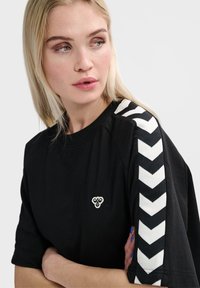Zwarte t-shirt met korte mouwen, voorzien van een wit chevronpatroon op de schouders en een klein embleem op de borst. Gemaakt van zacht materiaal.