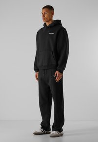 Haut à capuche noir avec poche avant et logo blanc, associé à un pantalon de survêtement noir assorti. Le modèle porte des baskets blanches et grises. Texture lisse.