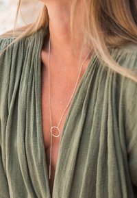 Long collier en argent avec pendentif circulaire et barre porté sur un haut en tissu texturé vert olive avec un col en V profond.