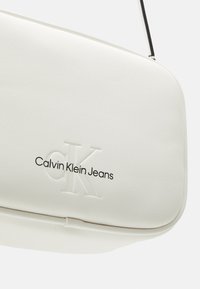 Biała skórzana torba na ramię z wypukłym logo "cK" oraz tekstem "Calvin Klein Jeans" w kolorze czarnym na przedniej części.