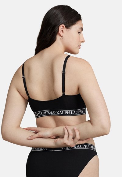 Bralette negro con canalé, con tirantes ajustables y una banda de logo blanca. La parte trasera presenta un diseño minimalista, que destaca por su tejido suave y detalles de costura.